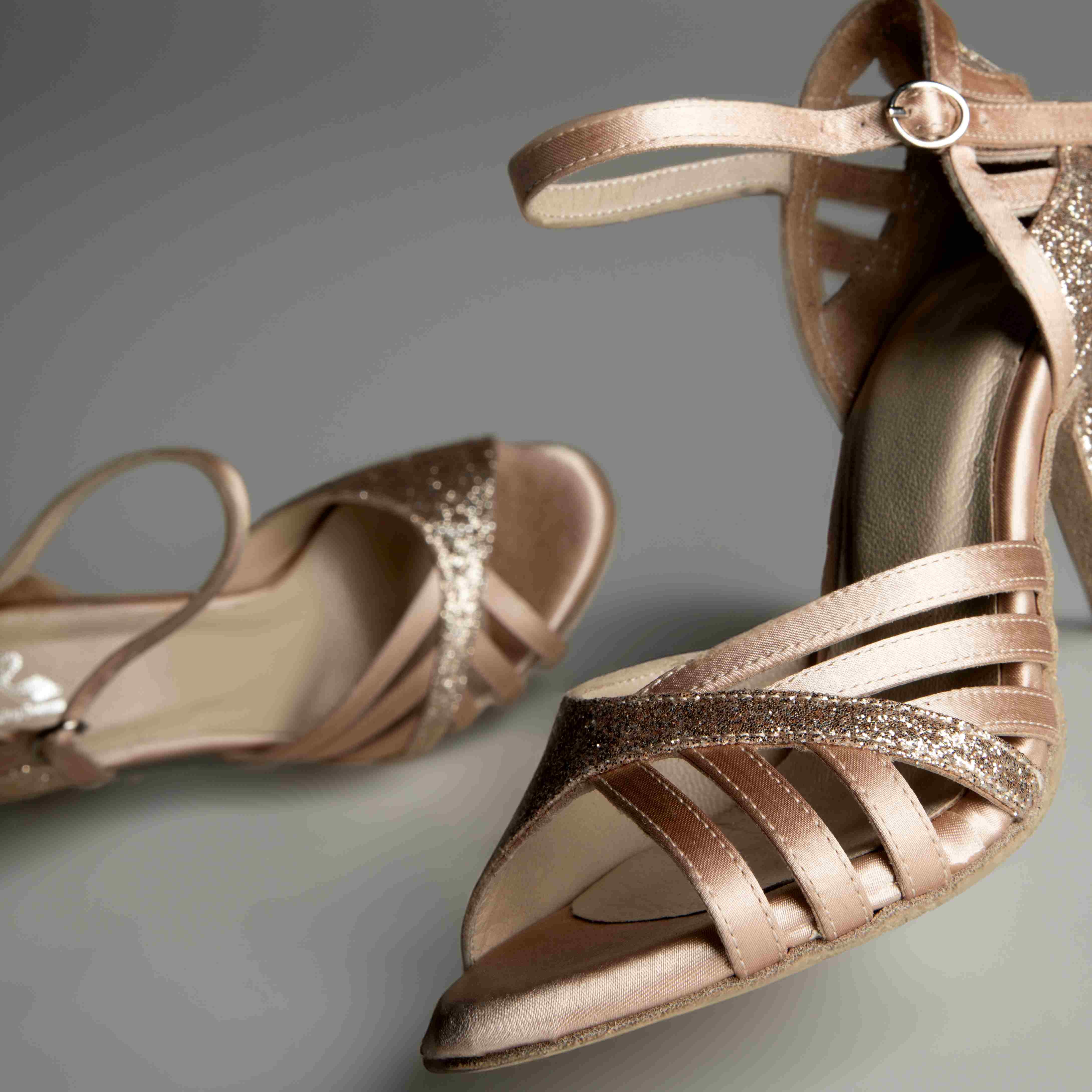 Goldene, glitzernde Riemchenschuhe mit hohen Absätzen, einer aufrecht und einer auf der Seite liegend, vor einem grauen Hintergrund.